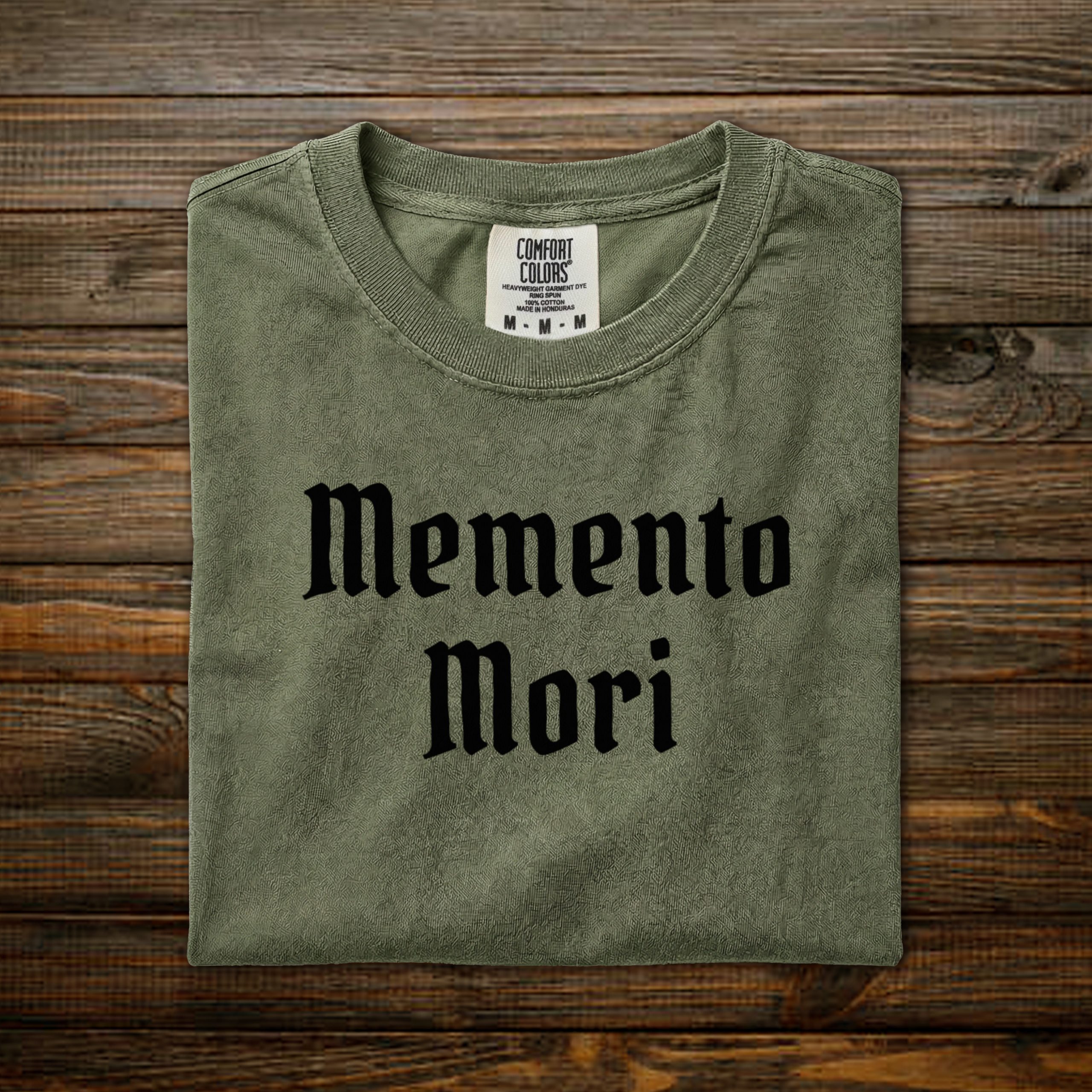 Memento Mori Comfort Colors tee with black old English lettering, folded on wood background