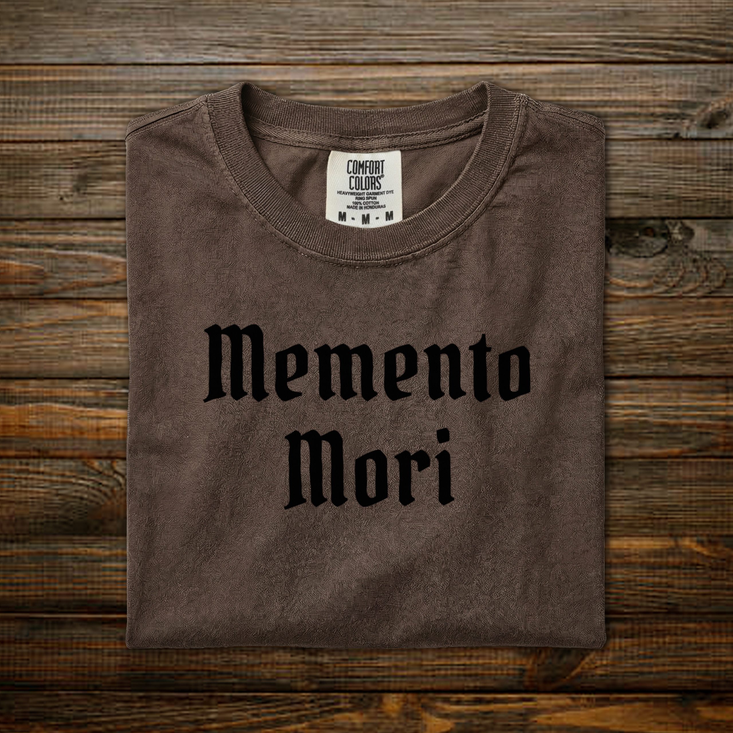 Memento Mori Comfort Colors tee with black old English lettering, folded on wood background