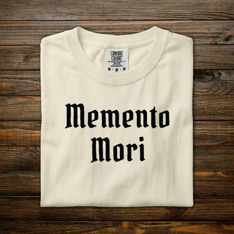 Memento Mori Comfort Colors tee with black old English lettering, folded on wood background