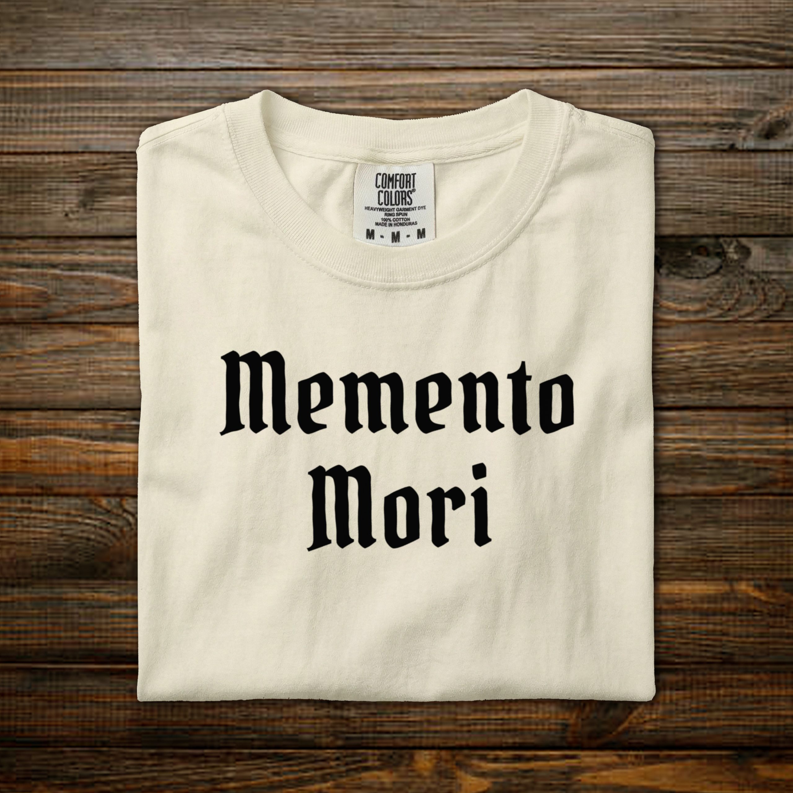 Memento Mori Comfort Colors tee with black old English lettering, folded on wood background