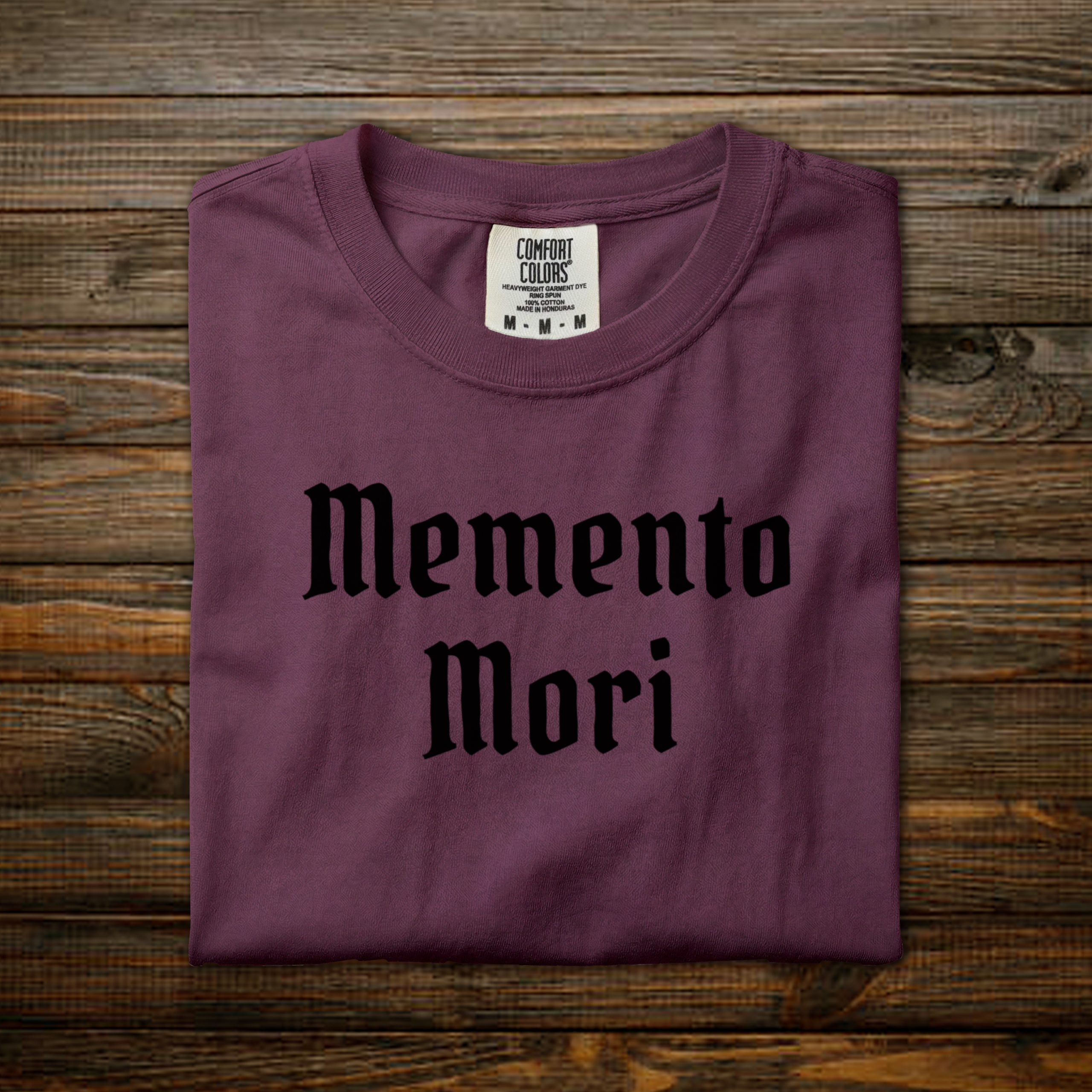 Memento Mori Comfort Colors tee with black old English lettering, folded on wood background