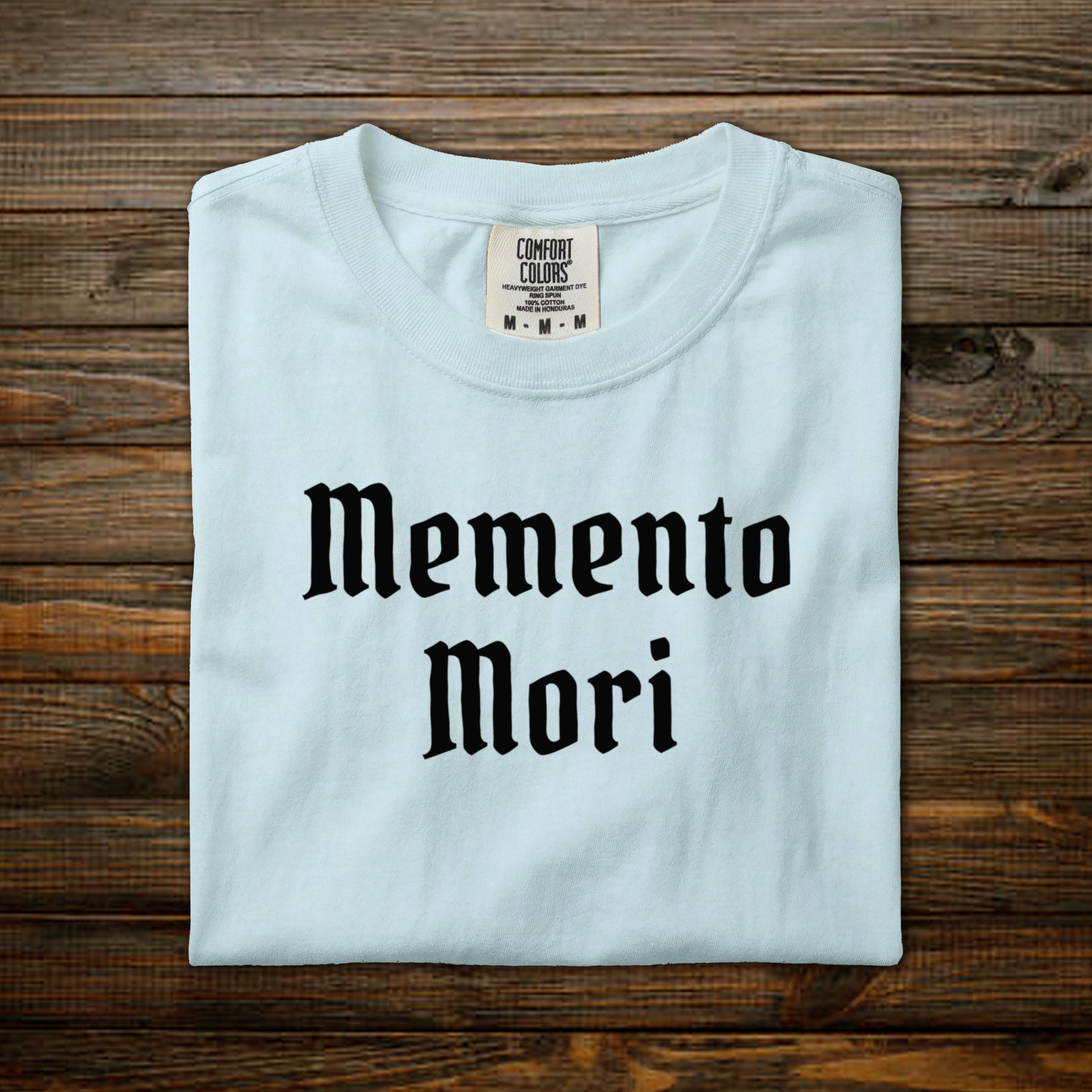 Memento Mori Comfort Colors tee with black old English lettering, folded on wood background