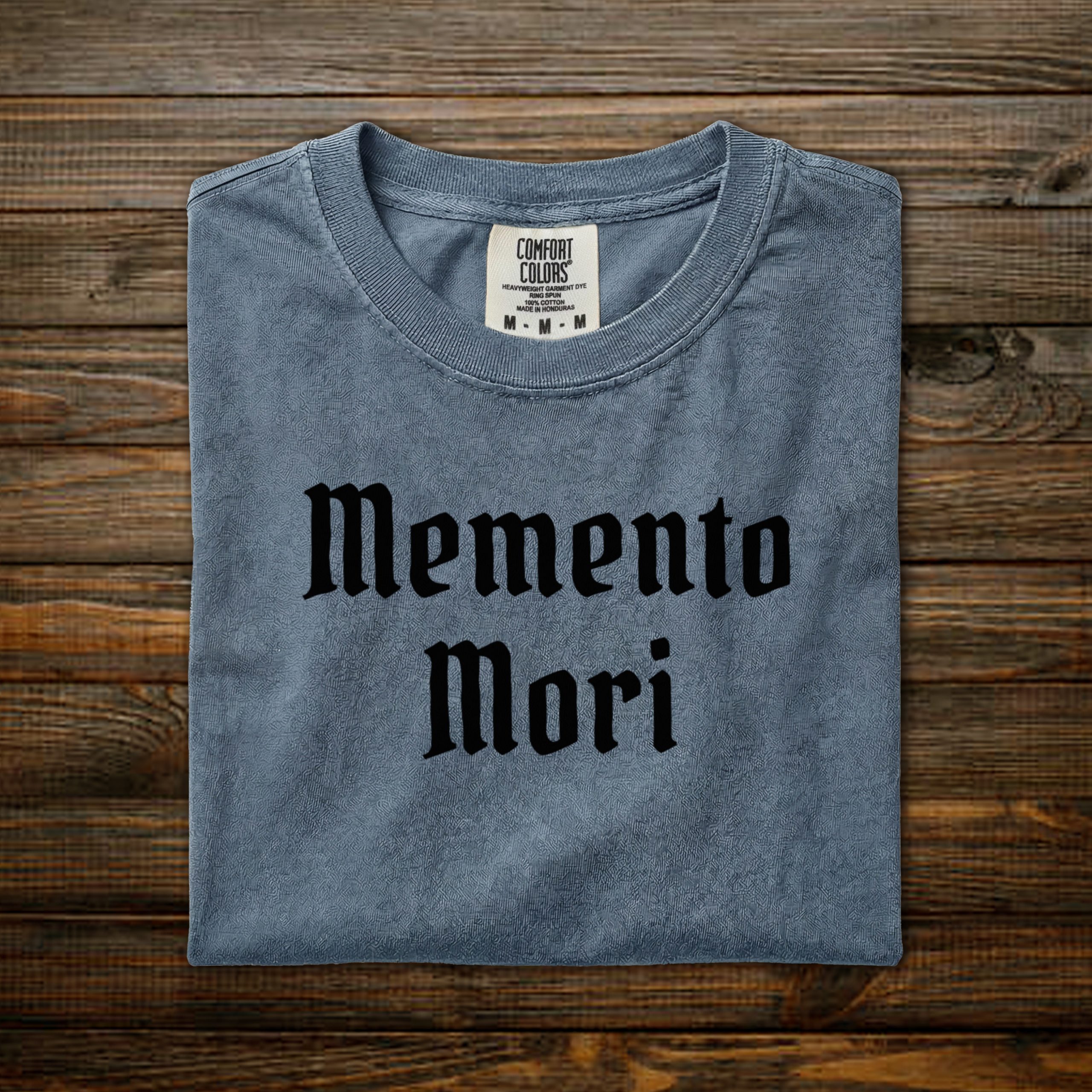 Memento Mori Comfort Colors tee with black old English lettering, folded on wood background
