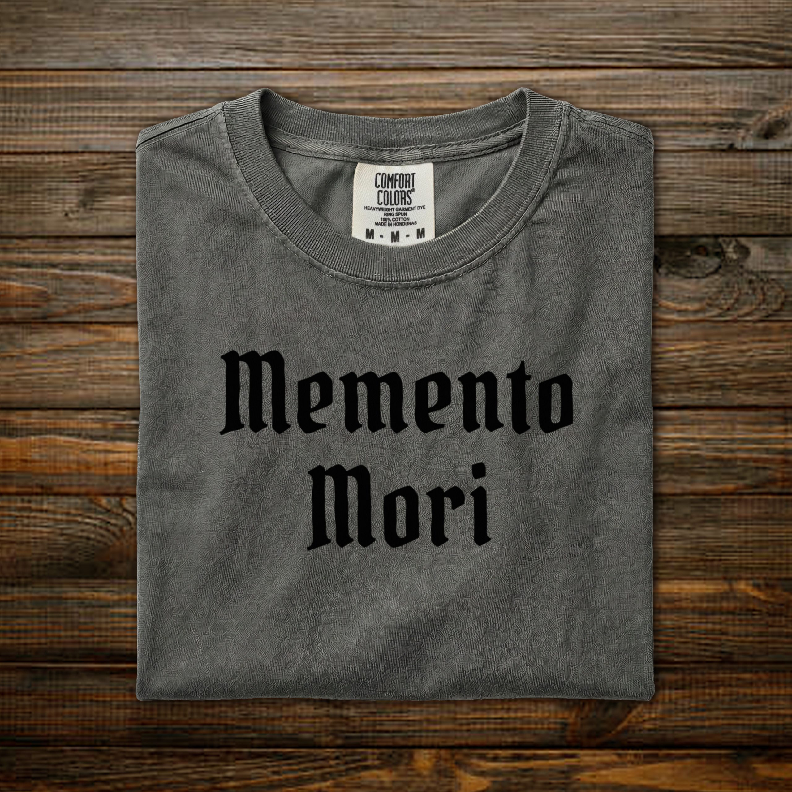 Memento Mori Comfort Colors tee with black old English lettering, folded on wood background